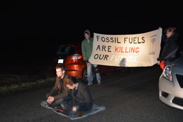 Alice Fine and Daniel Guering (seated) risk arrest with sign-holding activists standing behind them in the middle of Rocky Branch Road in Harrisburg, Ill. (Photo: James Anderson)