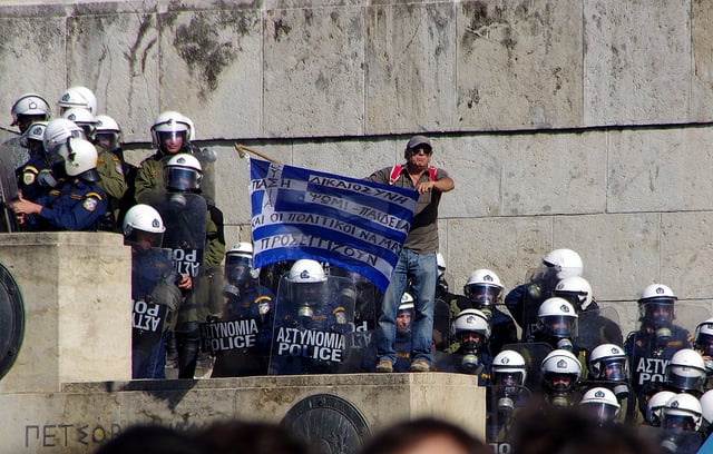 A protester climbs behind riot police to display a Greek flag bearing a populist message, in October 2011. (Photo: Eric Vernier)