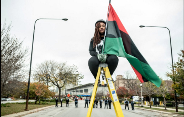 Cairá Lee Conner sits chained on top of a ladder, holding the Pan-African flag. (Photo: Sarah Jane Rhee)