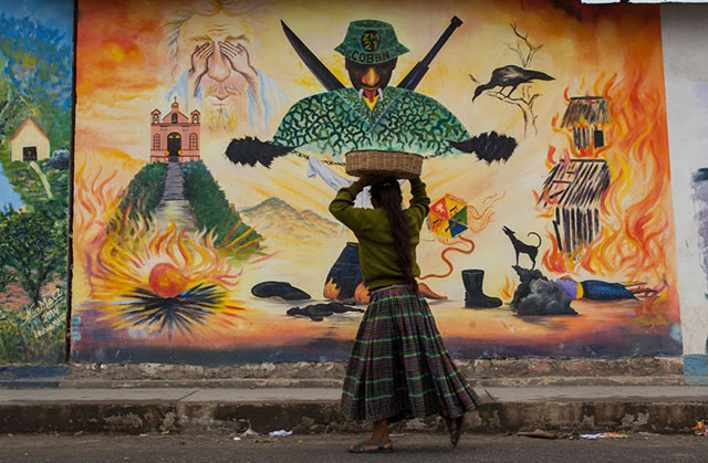 A young girl in San Cristobal Verapaz, Alta Verapaz, Guatemala passes in front of a mural depicting a soldier from Military Zone 21, and the military's scorched earth policy in Alta Verapaz. (Photo: Jeff Abbott)