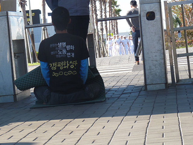 A protester is blocked by a security guard as he sits in silent protest outside the entry to the South Korean naval base on Jeju island. (Photo: Jon Letman)